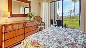 A bedroom with a large bed, a dresser, a mirror, and a view of a garden and ocean through a sliding glass door. - Peaceful Paradise! Spacious Condo, Full Kitchen, Wifi, Flat Screens-Kaha Lani 108 (LIHUE)
