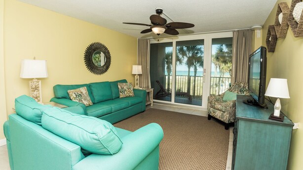 A living room with a teal sofa, a ceiling fan, a TV, and a view of the beach through the sliding glass door. - C106 Low Floor, Stunning sunset, Pool View, covered parking space (Panama City Beach)