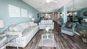 A spacious living room with a large sectional sofa, a smaller sofa, and a glass coffee table. There is a dining area with a table and chairs visible in the background.