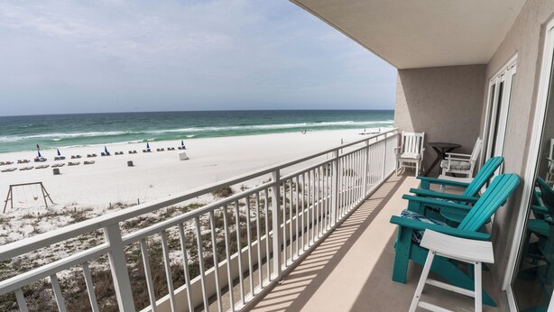 A balcony with a view of a beach and ocean. There are beach chairs and a table on the balcony.