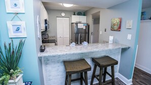 A kitchen with a marble countertop, two stools, a refrigerator, and a clock on the wall.