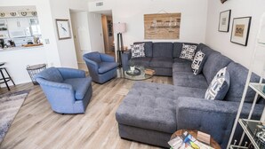 Una sala de estar moderna con un sofá seccional gris, dos sillones azules, una mesa de centro de vidrio y suelo de madera.