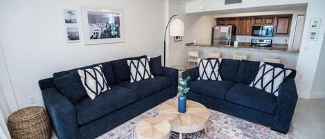 A living room with two dark sofas, a coffee table, and a patterned rug. There are framed pictures on the wall and a kitchen area with wooden cabinets and appliances.