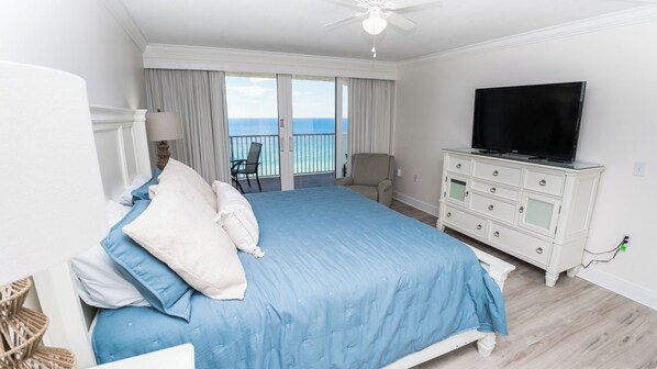 A hotel room with a large bed, a TV, a balcony with a view of the ocean, and a white dresser.