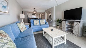 A living room with a blue sofa, a coffee table, a TV on a wooden stand, and a kitchen in the background. - D504 Gulf Front Views, 3 pools, Tennis (Panama City Beach)