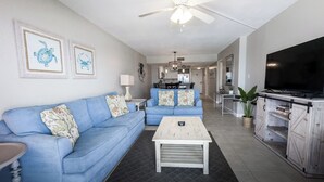 A living room with a blue sofa, a coffee table, and a TV on a wooden cabinet. - D504 Gulf Front Views, 3 pools, Tennis (Panama City Beach)