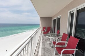 Een balkon met een glazen tafel en rode stoelen, uitkijkend op een strand met helder turquoise water.