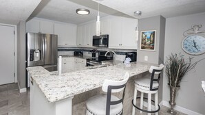 A modern kitchen with white cabinetry, stainless steel appliances, a marble countertop, and two bar stools. - D1005 Direct Gulf View, 3 Pools, Tennis and Pickle Ball (Panama City Beach)