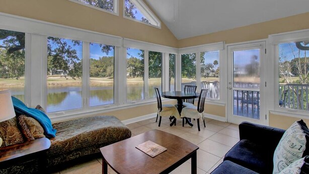 A sunroom with large windows overlooking a lake, a dining area with chairs, and a ceiling fan. - 15 Lake Village Lane by AvantStay | Golf Course & Lagoon Views (Isle Of Palms)
