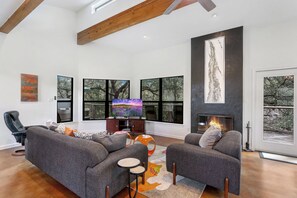 A modern living room with a fireplace, large windows, and a cozy seating area. - Secluded Wimberley Creekfront Home | Hot Tub, Fire Pit & Hill Country Views (Wimberley)