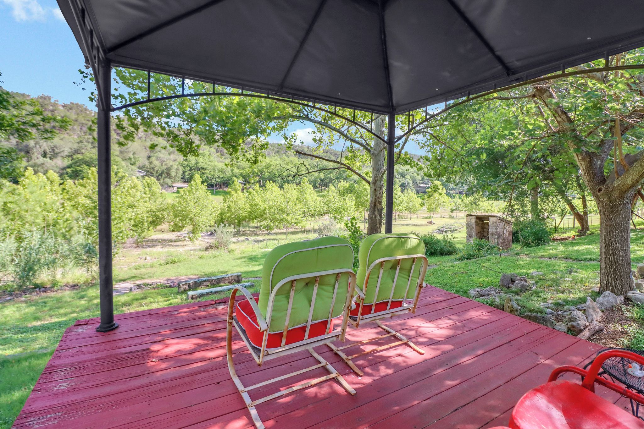 A covered outdoor seating area with two chairs and a table, surrounded by greenery and trees.