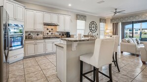 A modern kitchen with white cabinetry, a central island, and a dining area with a white sofa. - Fairway Pinehurst: Theater, Pool, Water Park, Golf View, Near Disney (Kissimmee)