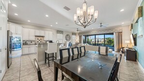 A spacious kitchen with white cabinets, a large island, and a dining area with a glass-top table and chairs. - Fairway Pinehurst: Theater, Pool, Water Park, Golf View, Near Disney (Kissimmee)