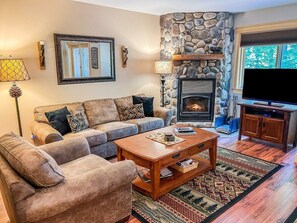 A living room with a stone fireplace, a sofa set, a coffee table, and a flat-screen TV. - Townhome D2 - Vacation Rental on 4th Lake (Inlet)