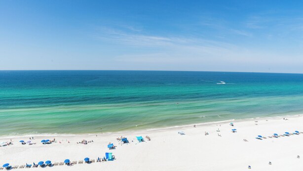 Ein Strand mit weißem Sand, klarem türkisblauem Wasser und einigen Sonnenschirmen und Liegestühlen.