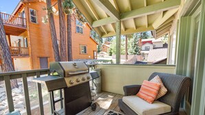 A covered outdoor area with a grill, a wicker sofa with cushions, and a view of a wooden house.