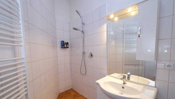 Une salle de bain avec des murs carrelés de carrelage blanc, une douche avec un pommeau de douche amovible, un lavabo avec un miroir et un porte-serviettes.