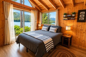 Une chambre à coucher avec un lit, des rideaux, une table de chevet, une lampe, une étagère avec des livres et des plantes, et une vue sur un lac et une forêt.