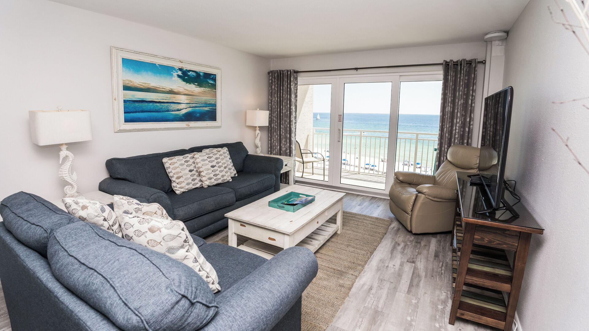 Ein Wohnzimmer mit Sofa, Sessel, Couchtisch und TV-Schrank. An der Wand hängt ein Gemälde und durch das Fenster ist der Strand zu sehen.
