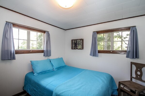 Un dormitorio con cama, dos ventanas con cortinas, cabecera de madera y un cuadro colgado en la pared.