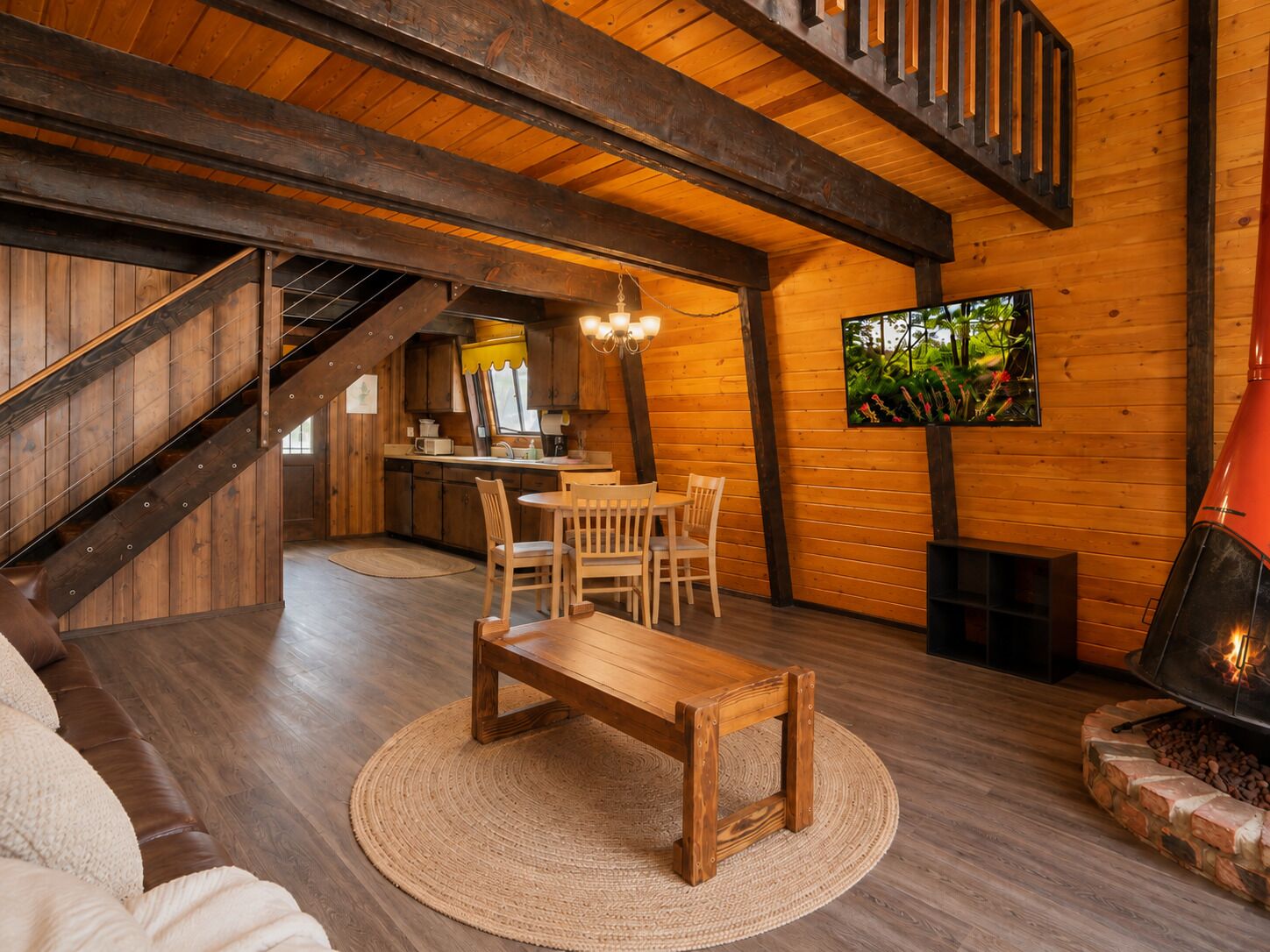 A cozy cabin interior with a wooden staircase, a dining area, a fireplace, and a kitchen.