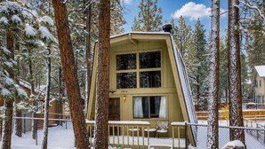 A snow-covered cabin with a unique sloped roof, surrounded by tall pine trees.