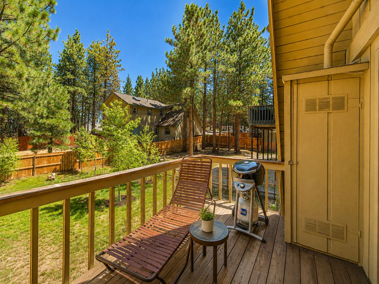 A wooden deck with a lounge chair and a grill, surrounded by trees and a house.