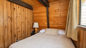 A wooden cabin room with a bed, a nightstand, a lamp, and a window with curtains. - Fanciful Winter's Lake - Portable AC, Walk to Lake, Close to Village, Fireplace, Barbecue (Big Bear Lake)