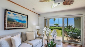 A living room with a sofa, a painting, a ceiling fan, and a view of the outdoors. - Kapaa Shore Ocean & Pool View Condo (Kapaa)