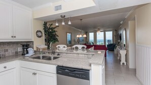 Una cocina moderna con encimeras de granito, lavavajillas y armarios blancos. Hay una zona de comedor con un sofá rojo y vistas al océano.