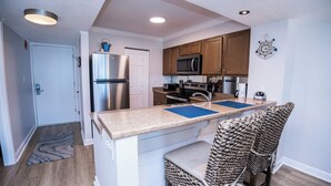 A compact kitchen with a refrigerator, microwave, and stove. There are two barstools and a small rug. - D303 Low Floor, Direct Gulf View, 3 pools (Panama City Beach)