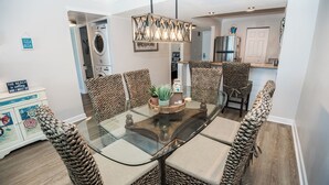 A dining area with a glass-top table surrounded by woven chairs, a hanging light fixture, and a kitchen in the background. - D303 Low Floor, Direct Gulf View, 3 pools (Panama City Beach)