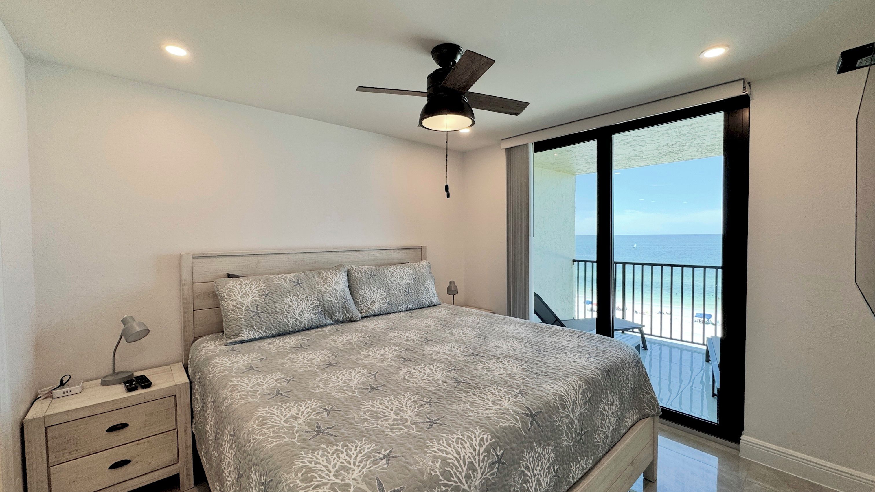Une chambre à coucher avec un lit, des tables de chevet, un ventilateur de plafond et une vue sur l’océan par une grande fenêtre.