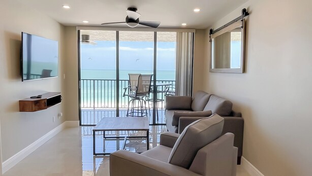 A modern living room with a balcony overlooking the sea, a flat-screen TV mounted on the wall, and a ceiling fan.