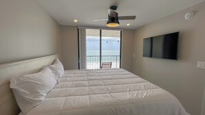 A bedroom with a bed, a ceiling fan, a TV, and a balcony with a view of the sea.