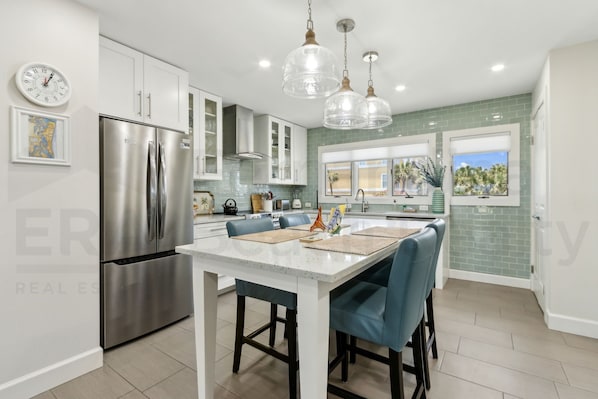 A modern kitchen with a stainless steel refrigerator, white cabinets, a central island with blue chairs, and pendant lighting.