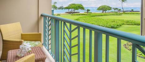 Un balcón con mobiliario de mimbre, vista a un césped cuidado y un paisaje tropical con árboles y el océano.