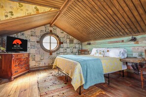 Un dormitorio acogedor en el ático con techo de madera, un ventanal redondo, una cama con colcha azul y amarilla, un aparador de madera y un televisor que muestra un logotipo.