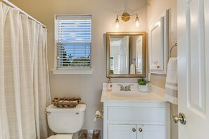 A bathroom with a toilet, a sink with a mirror, a window with blinds, and a shower curtain. - Beautiful views and the Crystal Clear waters of the Blanco River await you! (Wimberley)