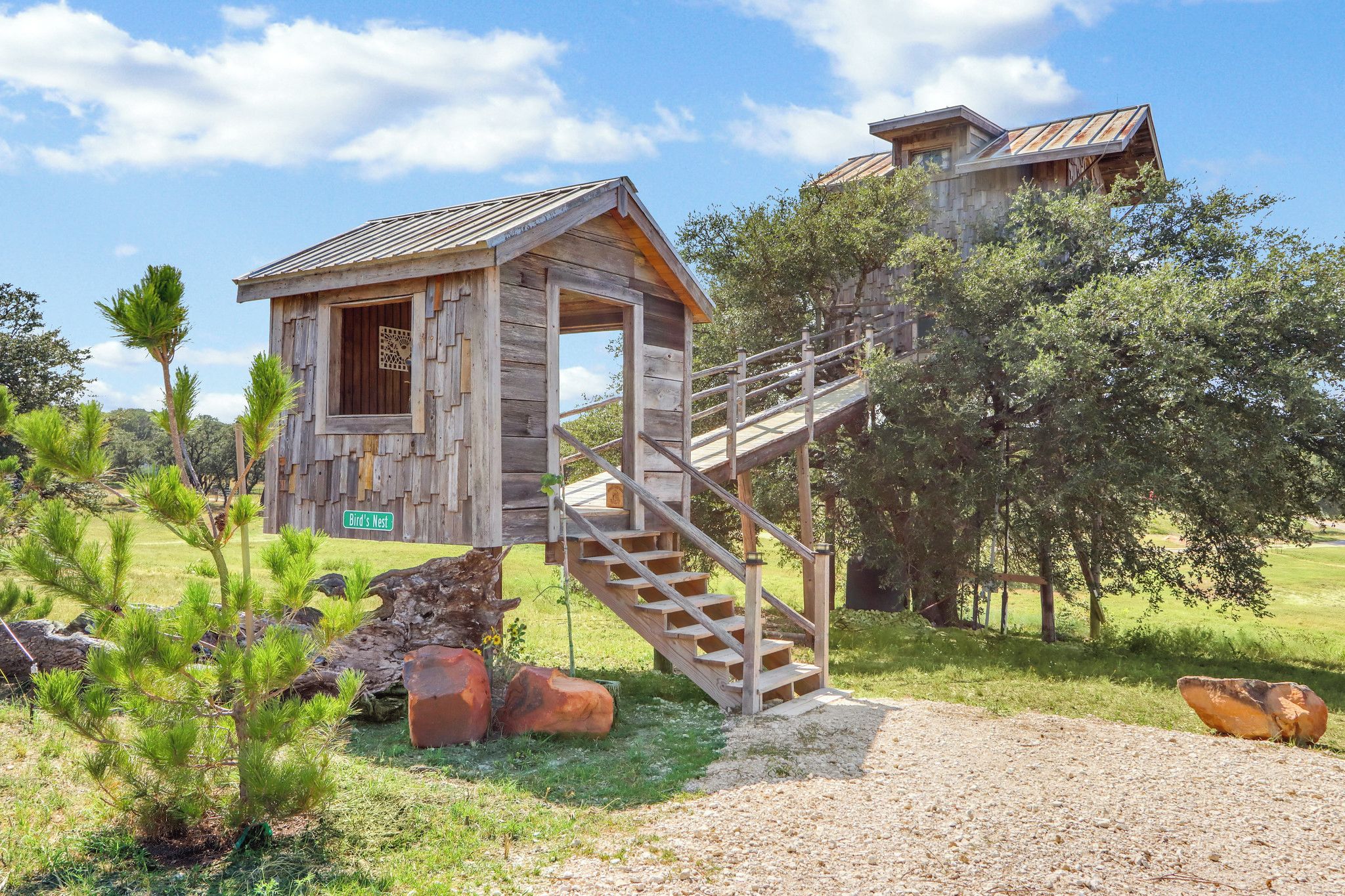 Une cabane en bois rustique, dotée d’un escalier menant à une mezzanine, entourée de verdure et d’arbres.