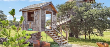 Une cabane en bois rustique, dotée d’un escalier menant à une mezzanine, entourée de verdure et d’arbres.