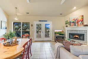 A living room with a fireplace, a dining area with a table and chairs, and a view through a door. - Beautiful views and the Crystal Clear waters of the Blanco River await you! (Wimberley)