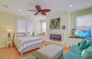 A bedroom with a bed, a ceiling fan, a fireplace, a television, and a window with blinds.