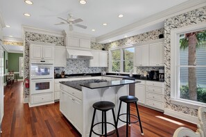 Une cuisine avec des meubles blancs, un îlot central doté de tabourets, un ventilateur de plafond et une fenêtre donnant sur un paysage verdoyant.