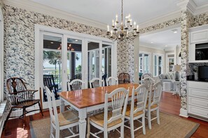 A dining area with a wooden table and white chairs, a chandelier, and a view through glass doors to the outside.