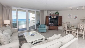 A spacious living room with a sofa, armchair, and coffee table. A television is on a wooden cabinet, and there is a dining area with chairs. The room has large sliding glass doors leading to a balcony with a view of the ocean. - E904 3 Gulf-front pools, Stunning Beach View, Tennis Courts (Panama City Beach)