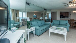 A spacious living room with a large mirror, two teal sofas, a white coffee table, and a dining area visible through the glass wall. - E1706 Gulf Front View, 3 Pools, Gated Condo (Panama City)