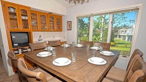 Un comedor con una mesa de madera dispuesta para seis personas, flanqueada por sillas de madera. A la izquierda se ve una cocina con armarios de madera y un televisor.