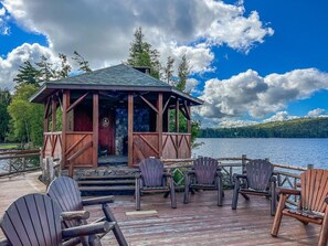 Eine Holzterrasse mit Adirondack-Stühlen und einem rustikalen Pavillon mit Blick auf einen See.