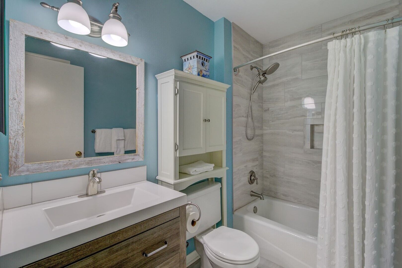 Une salle de bain avec un lavabo blanc, un miroir, des toilettes, une douche avec un rideau et un meuble de rangement.